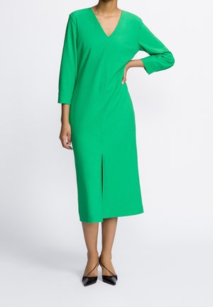 Donna che indossa un abito verde brillante al ginocchio con maniche a tre quarti e una scissione centrale frontale, abbinato a tacchi neri a punta.