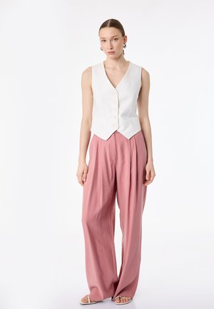 Mujer que lleva un chaleco blanco sin mangas con botones, pantalones sueltos rosas de talle alto y sandalias blancas, de pie sobre un fondo blanco liso.