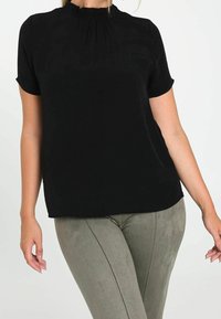 Une femme portant un haut noir à manches courtes avec un col froncé et un pantalon en daim gris ajusté, se tenant devant un fond uni.