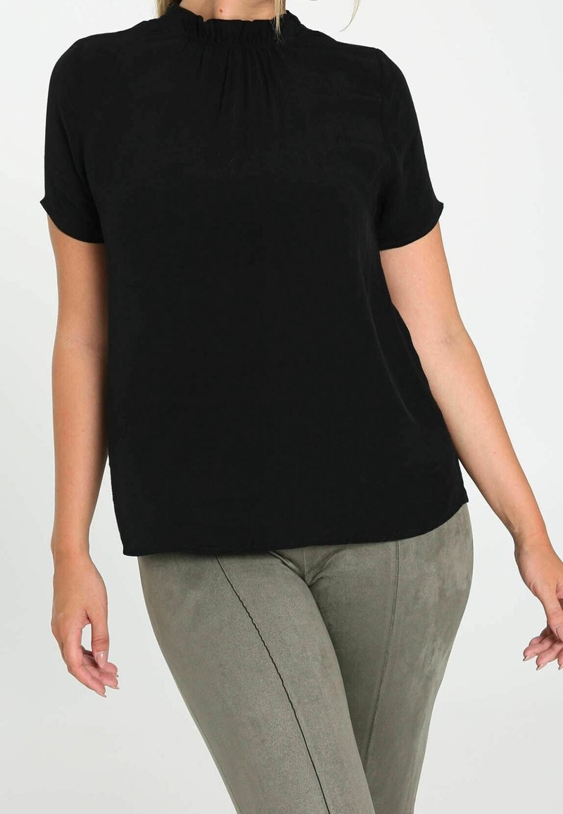 Une femme portant un haut noir à manches courtes avec un col froncé et un pantalon en daim gris ajusté, se tenant devant un fond uni.