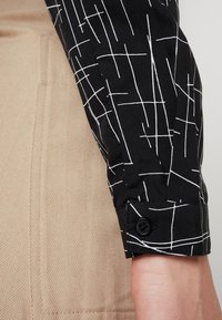 Svart skjorta med vit linjär mönstring, med knäppning vid cuff. Tyget har en slät textur mot en tan herringbone-bakgrund.