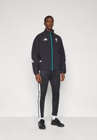 Μαύρο μπουφάν με φερμουάρ σε τιρκουάζ χρώμα και ροζ λεπτομέρειες, με λογότυπο της Liverpool FC. Συνδυασμένο με σκούρο αθλητικό παντελόνι και μαύρα αθλητικά παπούτσια.