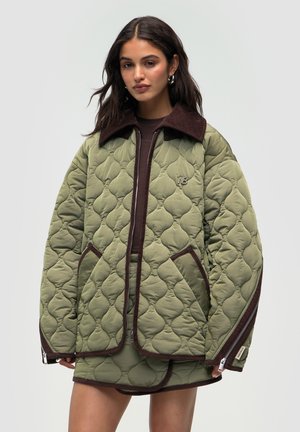 Jeune femme portant une veste en olive matelassée oversized avec des finitions marron foncé et une jupe matelassée assortie, se tenant devant un fond uni.