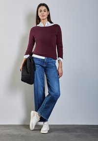 Pull bordeaux, chemise blanche à col en dessous, associé à un jean large bleu clair. Des baskets blanches et une veste matelassée noire sont tenues.