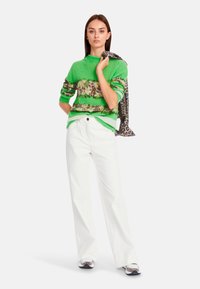 Grüner Strickpullover mit animalisch bedrucktem Bund, kombiniert mit weißen, weit geschnittenen Hosen. Das Model hält einen Schal mit Leopardenmuster. Strukturierte Stoffdetails sind sichtbar.