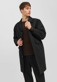 Svart ullrock med notched krage, enkel knappstängning och sidofickor, bärs över en brun texturerad tröja och mörka byxor.