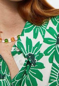 Camicetta floreale verde e bianca con collo a punta, caratterizzata da grandi motivi a foglia e dettagli con bottoni, arricchita da una collana di perline colorate.