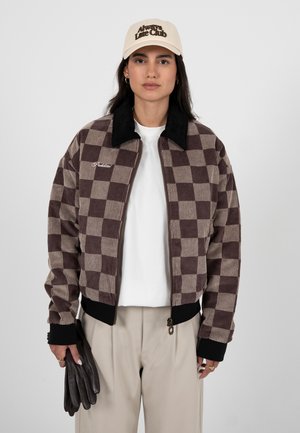 Frau trägt eine beige Mütze, braune karierte Jacke mit schwarzem Kragen, weißes Hemd, beige Hose und hält dunkelbraune Lederhandschuhe.