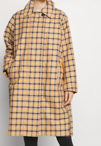 Długi płaszcz w kratę z żółtymi, brązowymi i purpurowymi wzorami, dużym kołnierzem i bocznymi kieszeniami. Wykonany z lekkiego materiału o luźnym kroju.