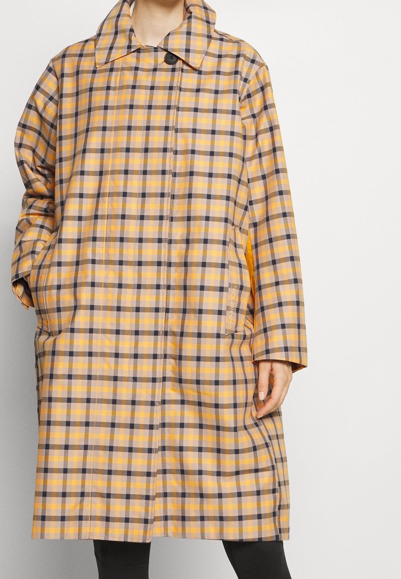 Długi płaszcz w kratę z żółtymi, brązowymi i purpurowymi wzorami, dużym kołnierzem i bocznymi kieszeniami. Wykonany z lekkiego materiału o luźnym kroju.