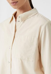 Camicia beige con bottoni, colletto, tessuto a trama, tasca frontale e bottoni a pressione argento. Presenta un logo ricamato discreto.