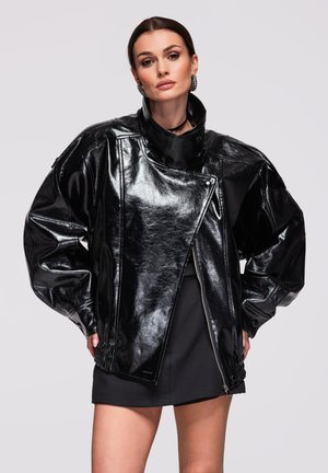 Kvinde iført oversized skinnende sort læderjakke med høj krave og sort mininederdel, poserer med hænderne på hofterne foran en ensfarvet baggrund.