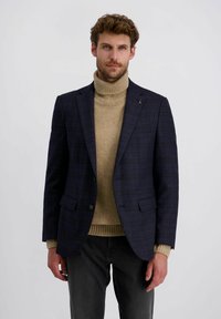 Mann in marineblazer mit Karomuster über beigem Rollkragenpullover und schwarzen Hosen, stehend vor einem schlichten weißen Hintergrund.