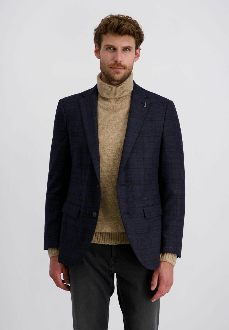 Mann in marineblazer mit Karomuster über beigem Rollkragenpullover und schwarzen Hosen, stehend vor einem schlichten weißen Hintergrund.