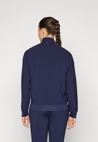 Navy jacket made of lightweight fabric features a high collar, long sleeves, and perforated panels on the sides for ventilation.