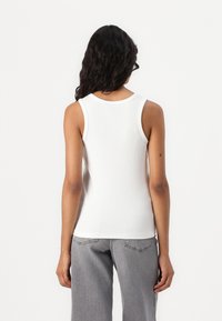 Camisola branca canelada com decote redondo e alças largas. Exibida pela parte de trás, combinada com calças cinzas.