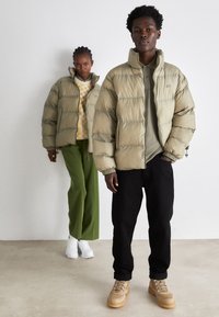 Chaqueta acolchada beige con secciones esponjadas, llevada sobre un suéter verde; pantalones negros y botas beige completan el look. La modelo está de pie frente a un fondo neutro.
