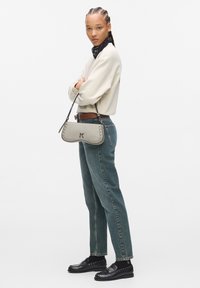 Borsa a tracolla in pelle grigia, con accenti di borchie e un logo. Il modello indossa un maglione crema e jeans blu sbiaditi.