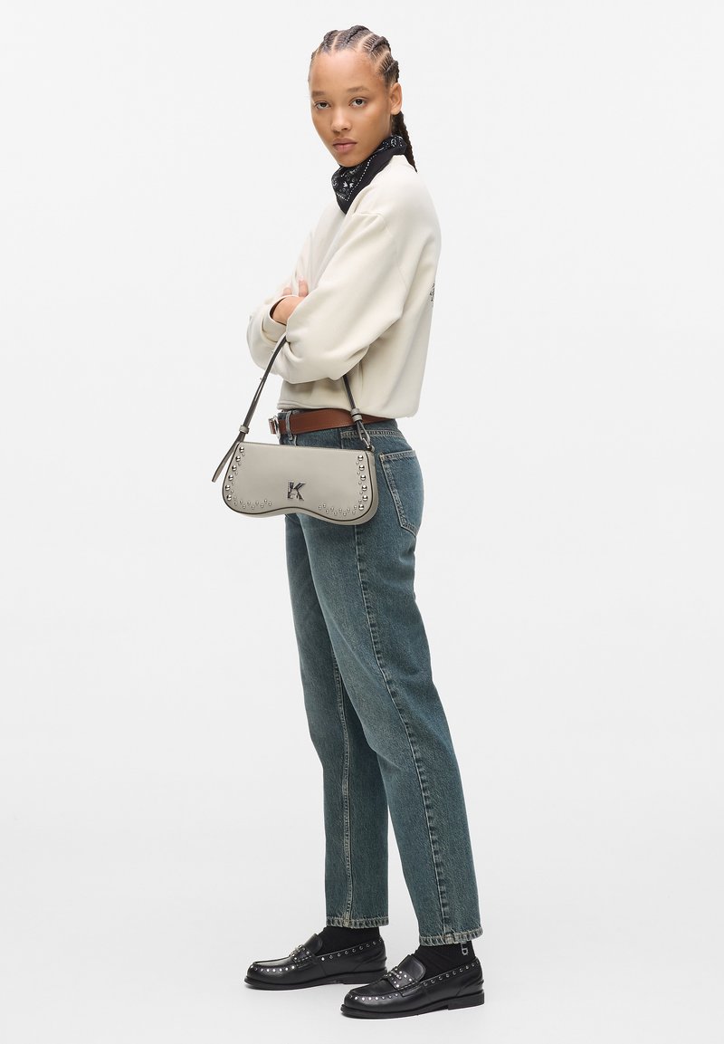 Borsa a tracolla in pelle grigia, con accenti di borchie e un logo. Il modello indossa un maglione crema e jeans blu sbiaditi.