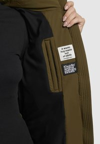 Grüne isolierte Jacke mit schwarzem Futter, ausgestattet mit zwei Etiketten: einem weißen mit Text und einem dunklen mit Logo. Sichtbares Reißverschlussfach.