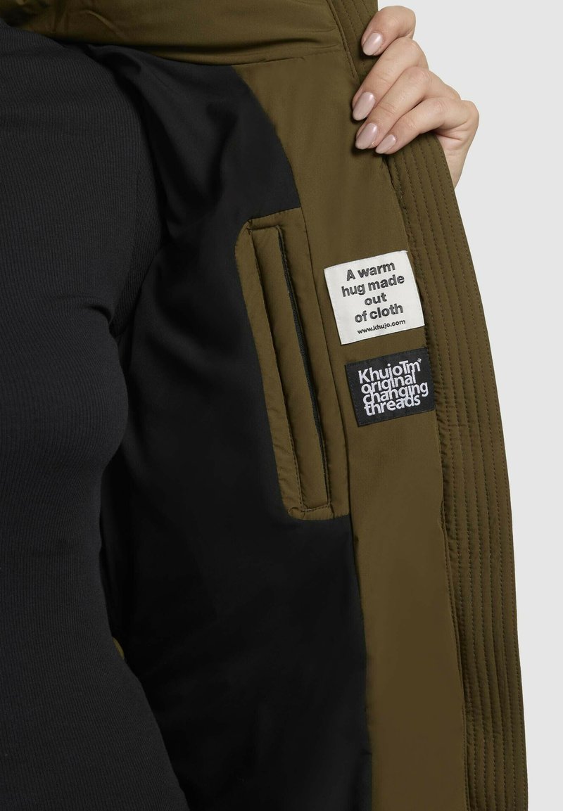 Giacca isolante verde con foderina nera, con due etichette: una bianca con testo e una scura con logo. Tasca con zip visibile.