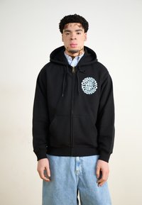 Juodasis zip-up džemperis su apvaliu šviesiai mėlynu logotipu kairėje pusėje, priekinėmis kišenėmis ir su raišteliu reguliuojamu kapučinu, vilkimas ant marškinėlių su apykakle.
