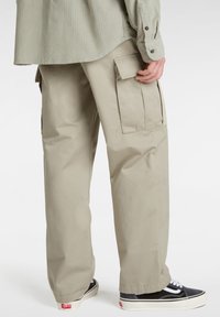 Beige cargo pants with large side pockets, loose fit, and lightweight fabric. Worn with black sneakers featuring white laces and a rubber sole.