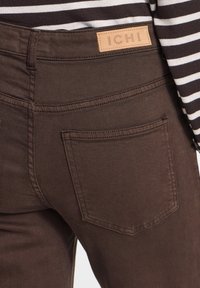 Pantalon marron avec poche arrière et étiquette de marque en cuir fauve "ICHI", porté avec un haut rayé noir et blanc.