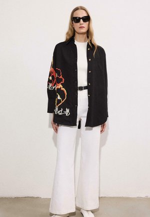 Γυναίκα που φοράει μαύρο πουκάμισο με κουμπιά, κεντημένο με κόκκινα και κίτρινα λουλούδια, λευκή μπλούζα, λευκό φαρδύ παντελόνι, μαύρη ζώνη, λευκά παπούτσια και γυαλιά ηλίου.