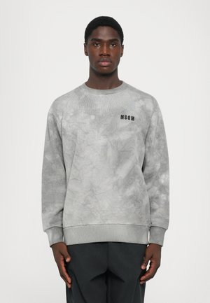 En ung mann står med fronten mot kameraet, iført en grå tie-dye MSGM genser og svarte bukser foran en enkel hvit bakgrunn.