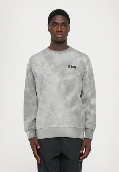 Un jeune homme se tenant de face, portant un sweat à capuche gris tie-dye de MSGM et un pantalon noir, devant un fond blanc uni.