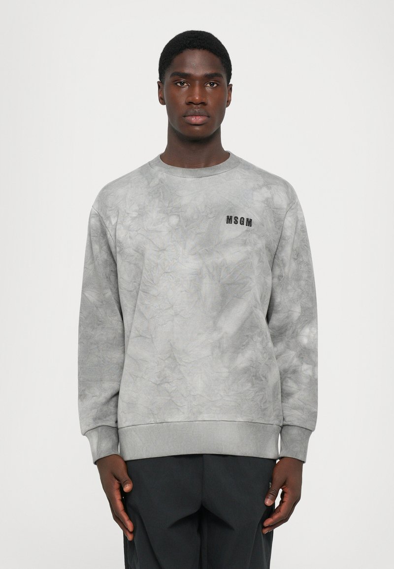Junger Mann, der mit einem grauen Batik-MSGM-Sweatshirt und schwarzen Hosen in einer frontal Blickrichtung vor einem schlichten weißen Hintergrund steht.