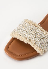Sandale en cuir marron à glissière avec une large sangle ornée d'un tissu crème texturé et de petites perles blanches, agrémentée de fils dorés.