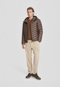 Giacca a vento marrone con design trapuntato, cappuccio attaccato e tasche con zip. Indossata sopra un top marrone, abbinata a pantaloni cargo beige e sneaker marroni.