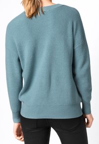 Femme portant un pull bleu ample en tricot et un pantalon noir, vue de dos avec les cheveux attachés en queue de cheval basse.