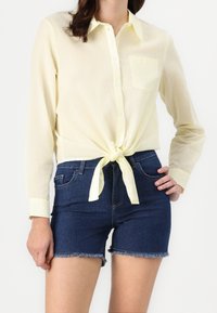 Chemise boutonnée jaune clair avec un nœud à l'avant, associée à un short en denim bleu foncé avec un ourlet effiloché.