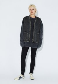 Černá denimová vesta se dvěma předními kapsami, přes tmavě šedou oversized košili. Kombinováno s černými úzkými džíny a bílými teniskami.