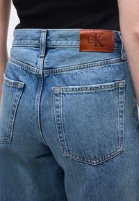 Εγγύς λήψη προσώπου που φοράει ανοιχτό μπλε ψηλόμεσο τζιν με μεγάλες πίσω τσέπες και καφέ δερμάτινο έμβλημα Calvin Klein στη μέση.