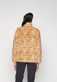 Veste en fausse fourrure beige avec motif à carreaux blancs, col haut et épaules tombantes. Texture douce et coupe décontractée. Vue de dos présentée.