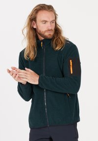 Mörkgrön fleecejacka med hög krage, framzip och orange accentdragkedjor. Har en svart patchficka på vänster arm.