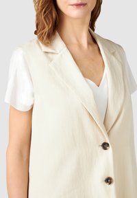 Světle béžová vesta se strukturovaným designem, klopami do tvaru písmene V, se třemi knoflíky a uvolněným střihem přes bílou košili s krátkým rukávem.