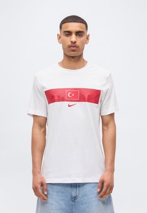 Mladý muž oblečený v bielom tričku s červenou tureckou vlajkou a logom Nike, v kombinácii so svetlomodrými džínsovými šortkami, stojaci pred jednoduchým pozadím.