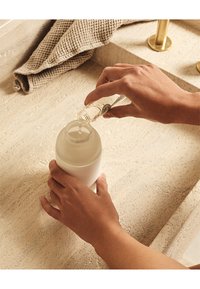 Bottiglia d'acqua in vetro satinato a forma arrotondata, tenuta in una mano, mentre si versa del liquido da una bottiglia trasparente all'interno, su una superficie testurizzata.