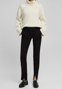 Krämfärgad stickad turtleneck-sweater med ribbad textur, i kombination med svarta skräddarsydda byxor med sidoflikar och svarta loafers.
