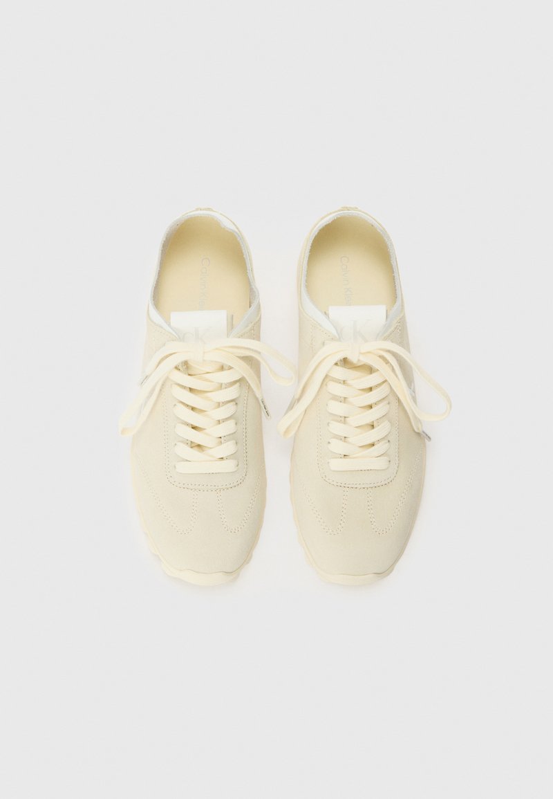 Zapatillas de deporte en un suave color beige claro, con parte superior de ante texturizado, puntera redondeada y suela de goma. Presentan cordones planos y un branding minimalista en la lengüeta.