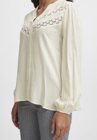 Blouse crème clair avec une finition texturée, comportant un panneau en dentelle sur la poitrine et des manches longues froncées. Design à boutons sur le devant.