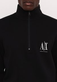 Personne portant un pull-over Armani Exchange noir à fermeture éclair partielle avec le logo blanc "A|X" sur la poitrine gauche et un col zippé.