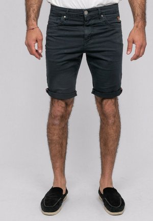 Homme portant un short en denim noir retroussé, une chemise blanche, des chaussures noires sans lacets, debout devant un fond clair uni.
