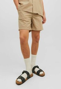 Personne portant un short beige texturé, des chaussettes blanches et des sandales à boucle noires, sur un fond clair uniforme.