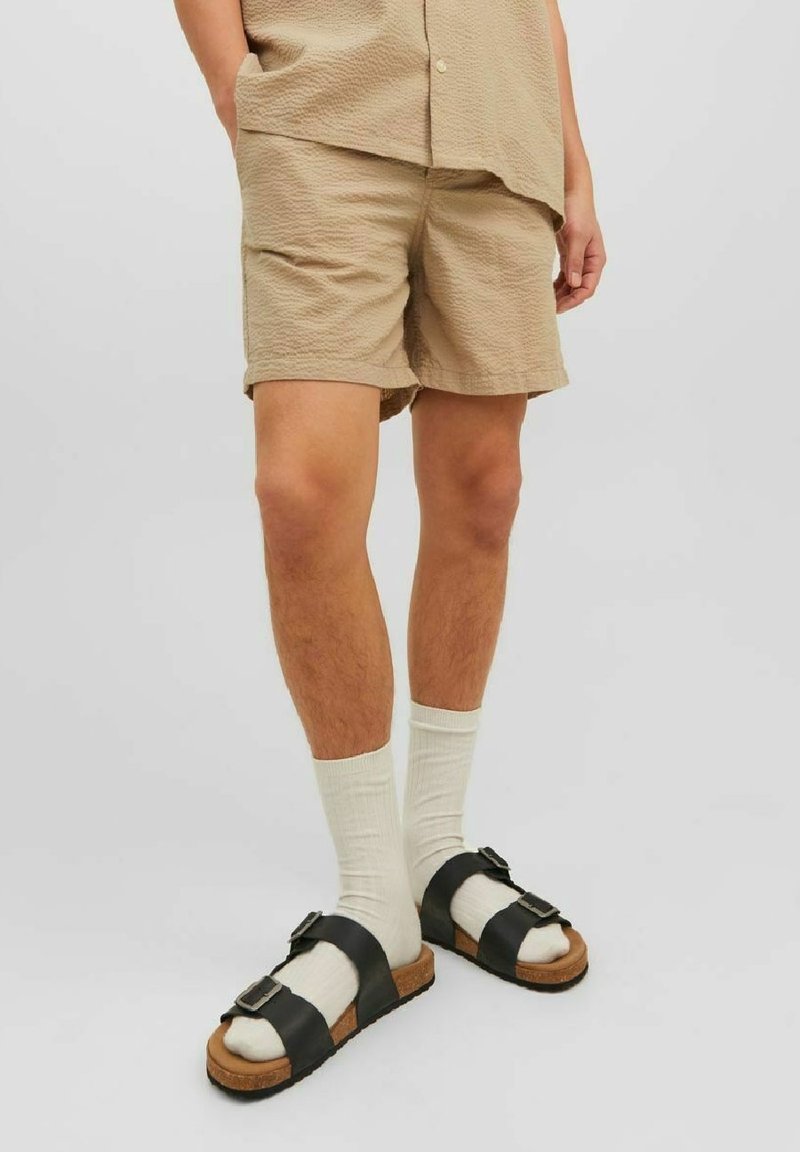 Personne portant un short beige texturé, des chaussettes blanches et des sandales à boucle noires, sur un fond clair uniforme.
